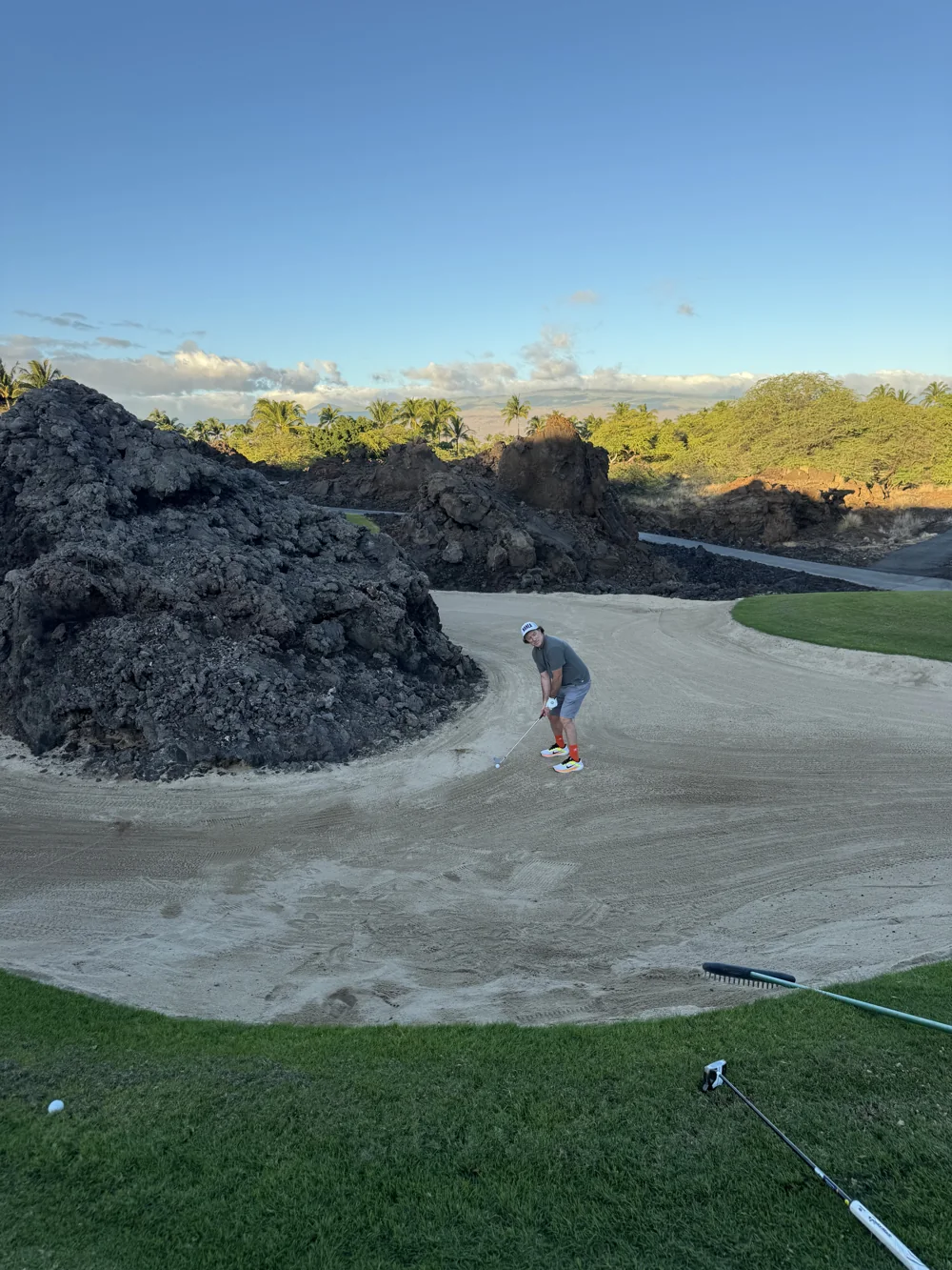 A golfer playing from a bunker beside dark rocks.